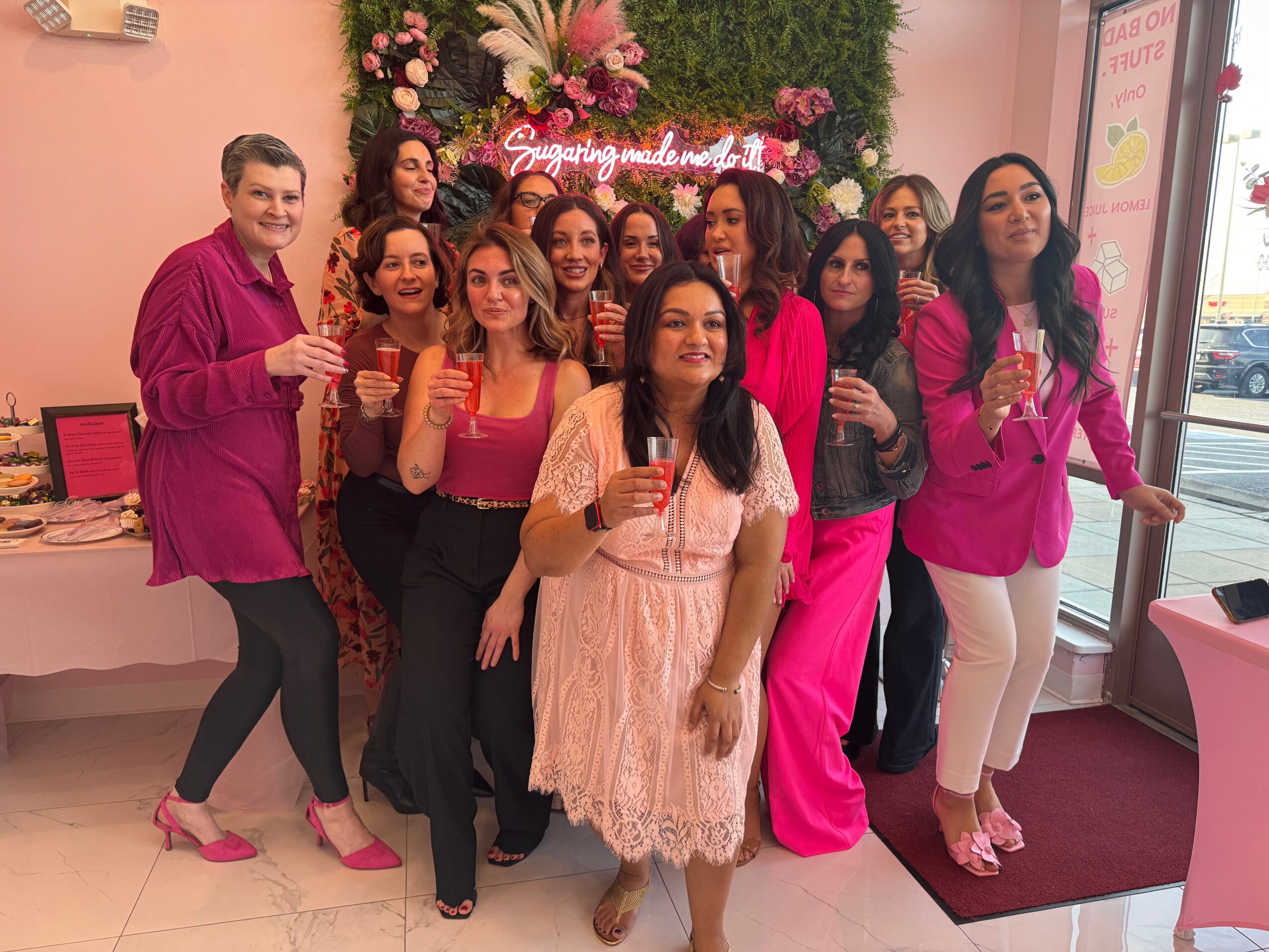 Group of women celebrating with drinks in front of floral decoration and a sign, indoors with pink decor.
