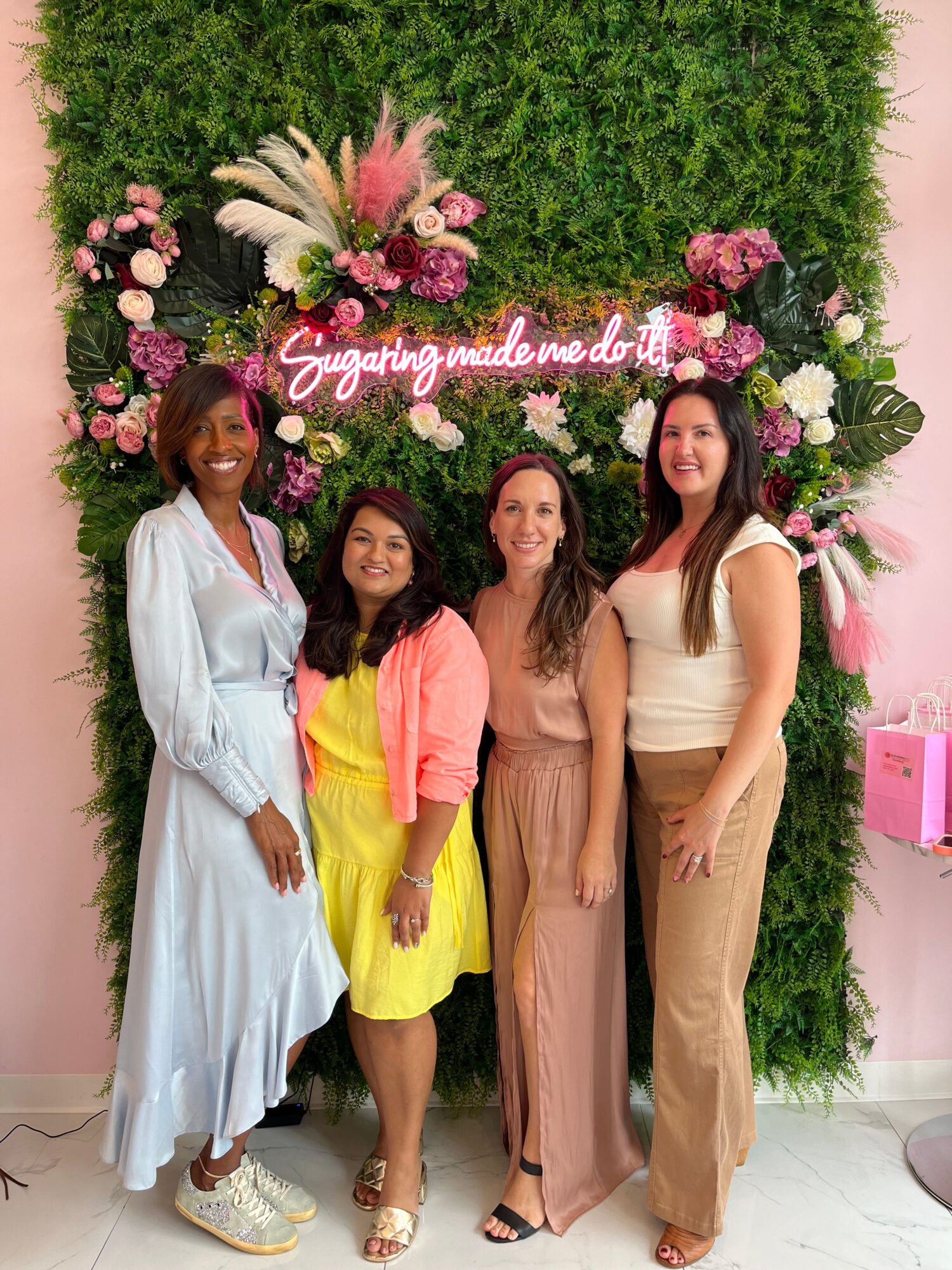 Four women standing in front of a floral backdrop with pink and white flowers, smiling, with greenery behind them.