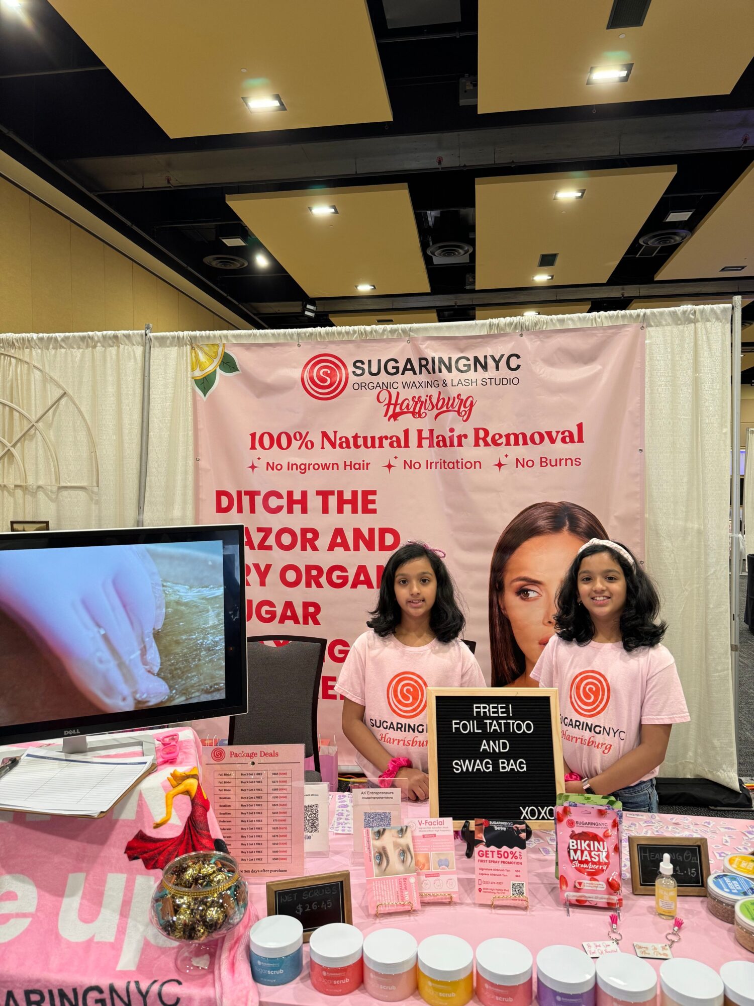 Two women standing behind a table with skincare products, promotional banner in the background, and a monitor displaying skincare images.