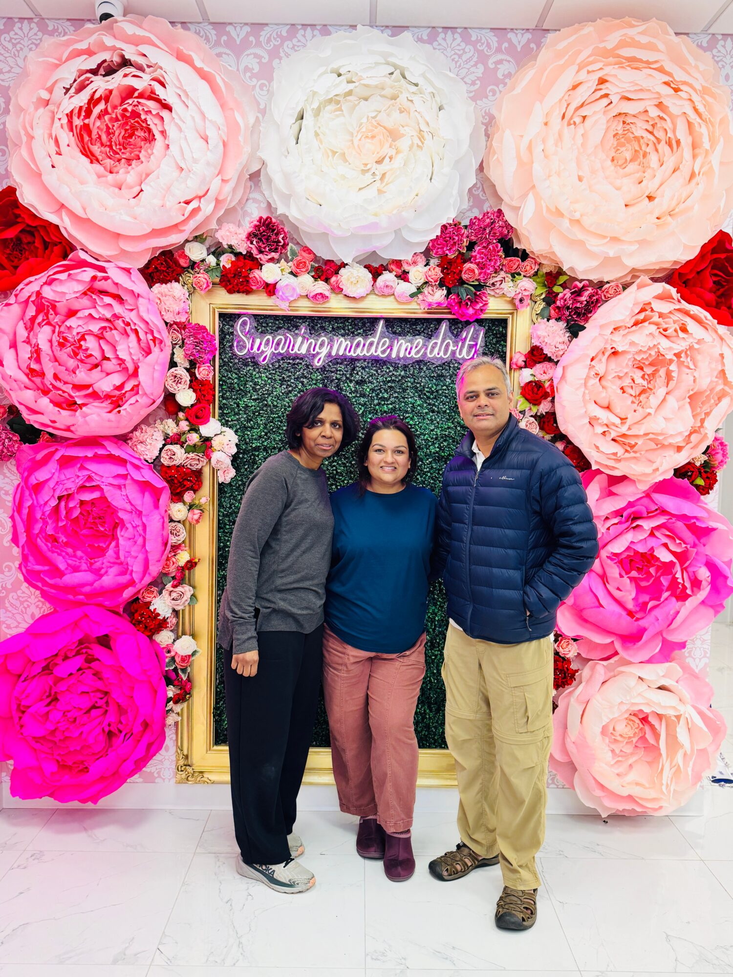 Three people stand in front of a floral backdrop with large pink, white, and peach paper flowers and a sign that reads 'sugaringsmademedoit'.