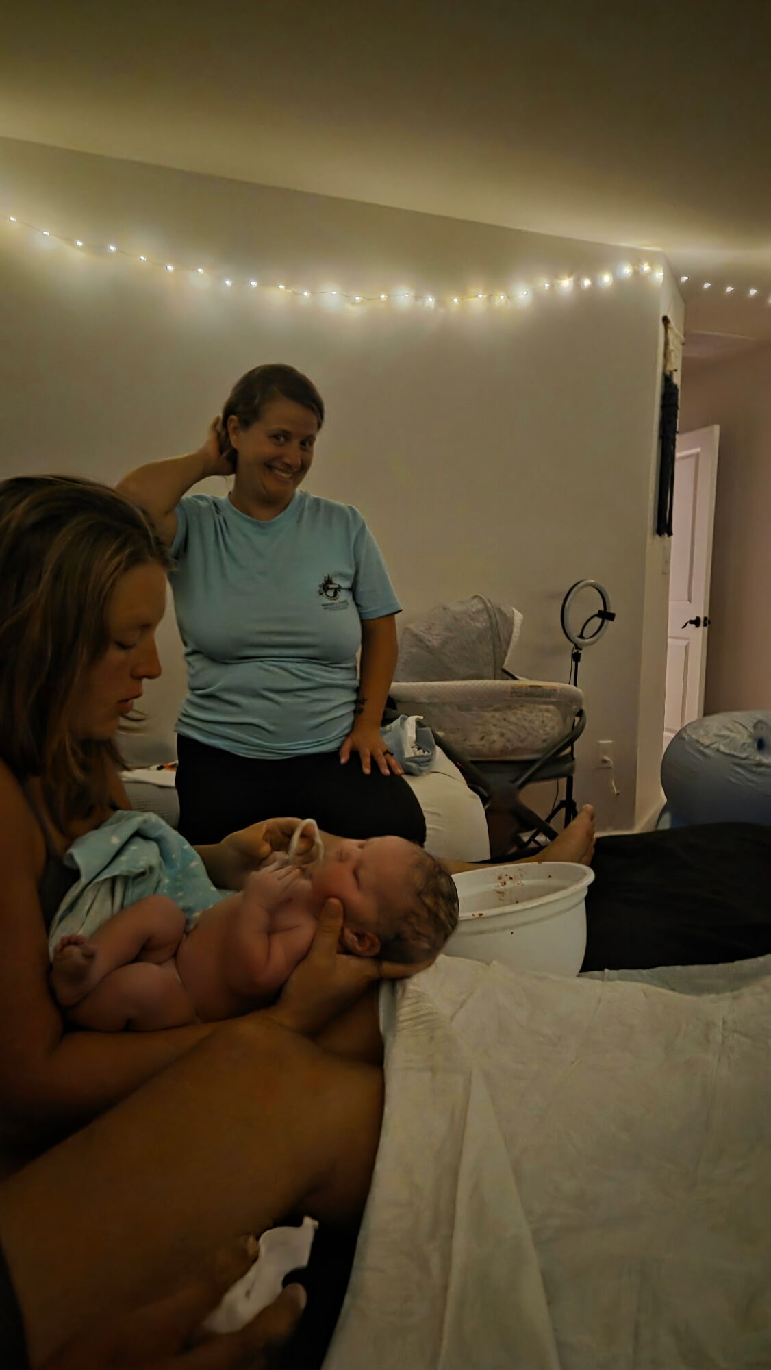 Person holding a baby, two women in the background, string lights on the ceiling, cozy indoor setting.
