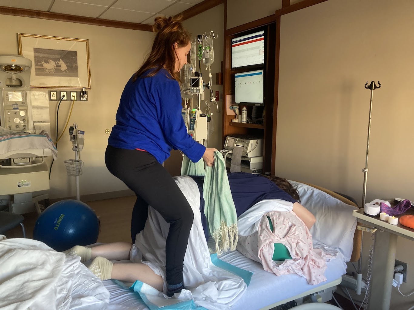 A woman in blue shirt and black pants stands beside a hospital bed with a patient, medical equipment visible in the background.
