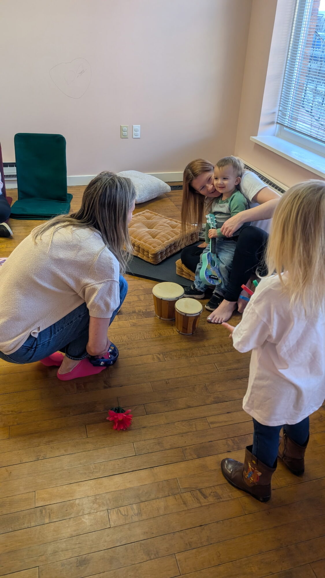 Adult and children playing a game with wooden cups on a wooden floor in a room with a window, a cushion, and a green chair.