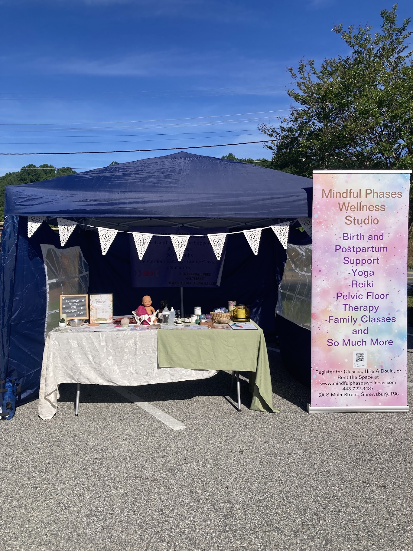 Outdoor wellness studio setup with a table under a canopy, decorated with bunting, and a sign listing services.
