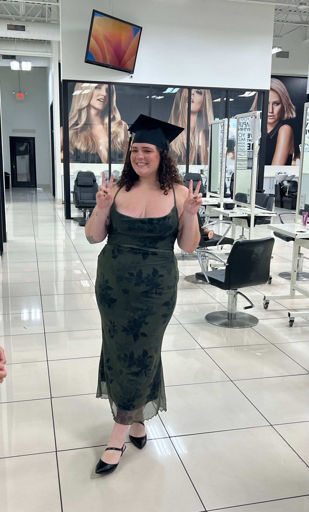 Woman in graduation cap and gown smiling and making peace signs in a store with mirrors and chairs