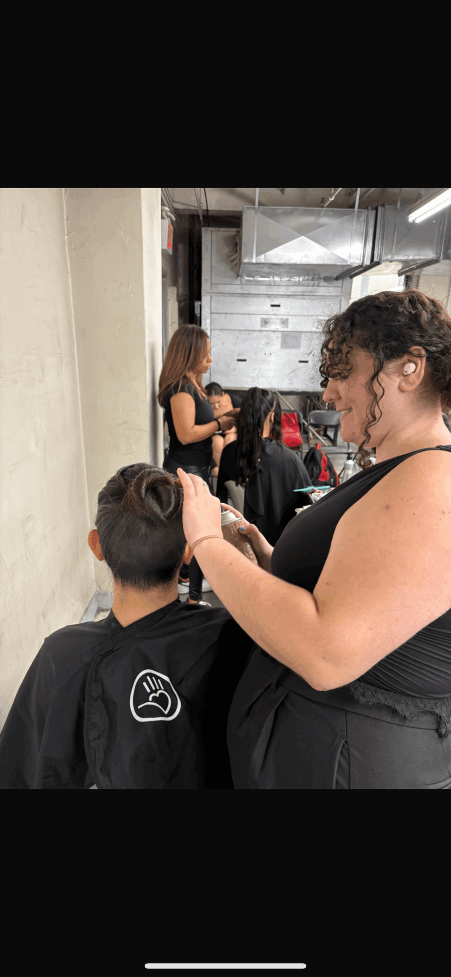 A woman with curly hair and glasses adjusts a person's hair in a busy indoor setting with people in the background.