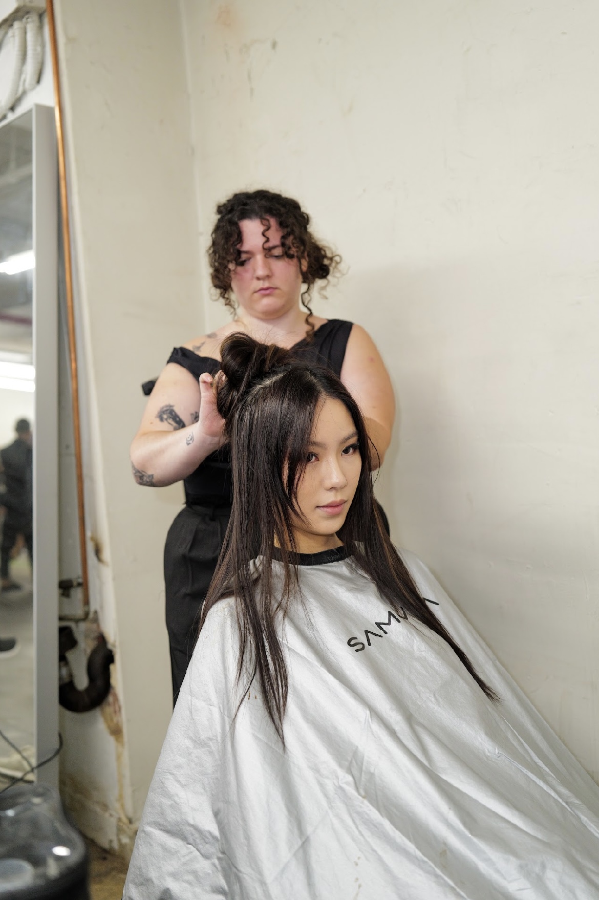 Hairdresser styles a woman's long hair in a salon, with a mirror and salon tools visible. Woman wears a cape.