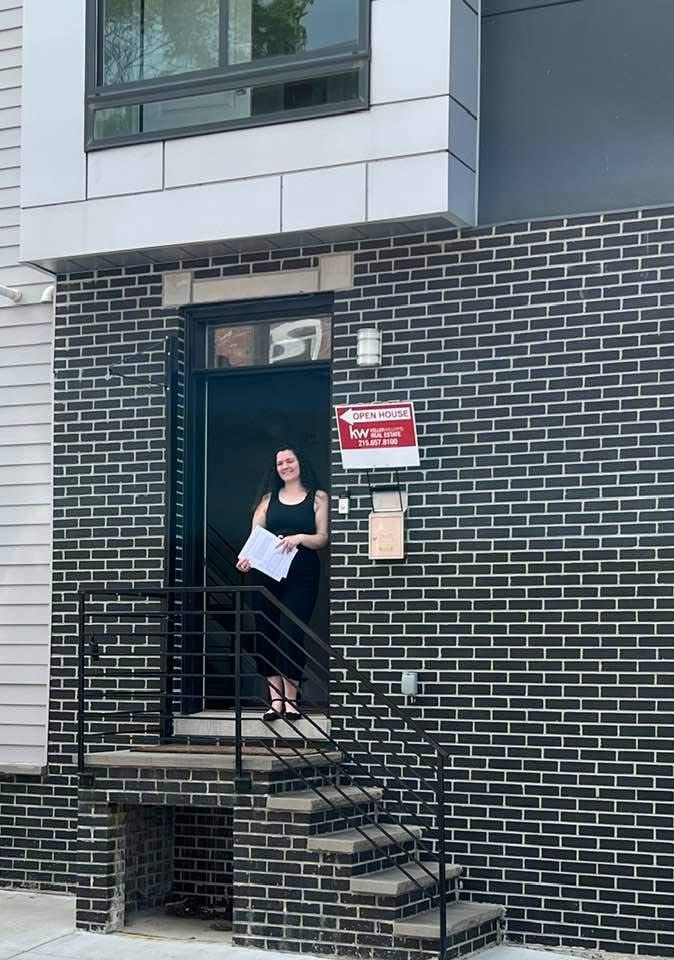 Woman standing on stairs outside a brick building holding papers, with a sign on the door.