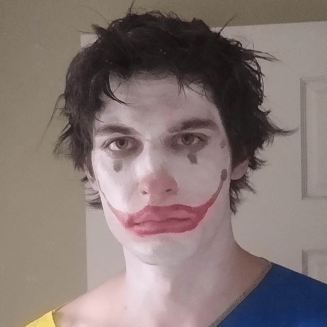 Person with clown makeup, messy dark hair, wearing a blue shirt, standing indoors near a door.