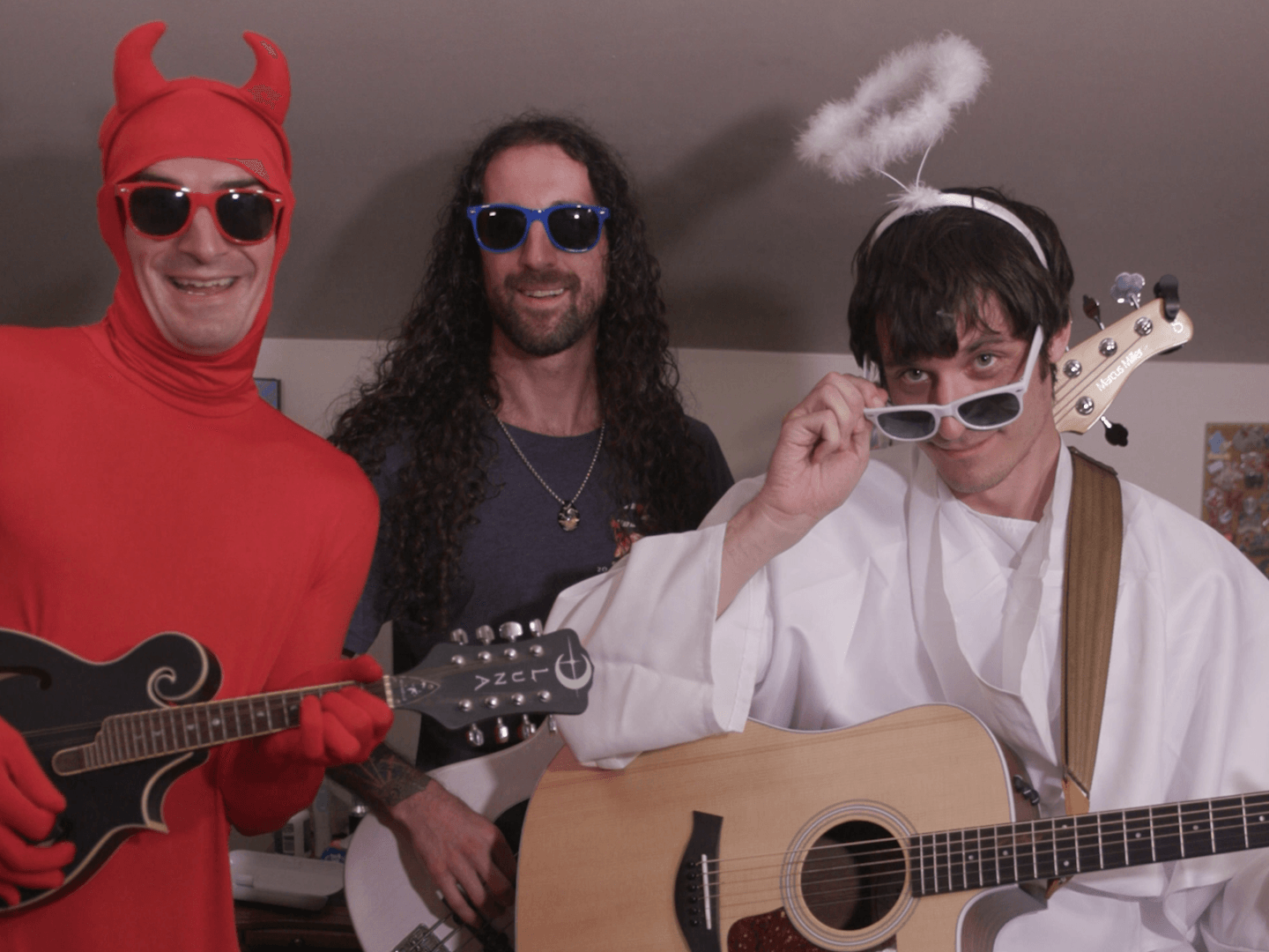 Three people dressed in costumes, one with devil horns and sunglasses, one with long curly hair and sunglasses, and one dressed as an angel with a halo and sunglasses, holding guitars.
