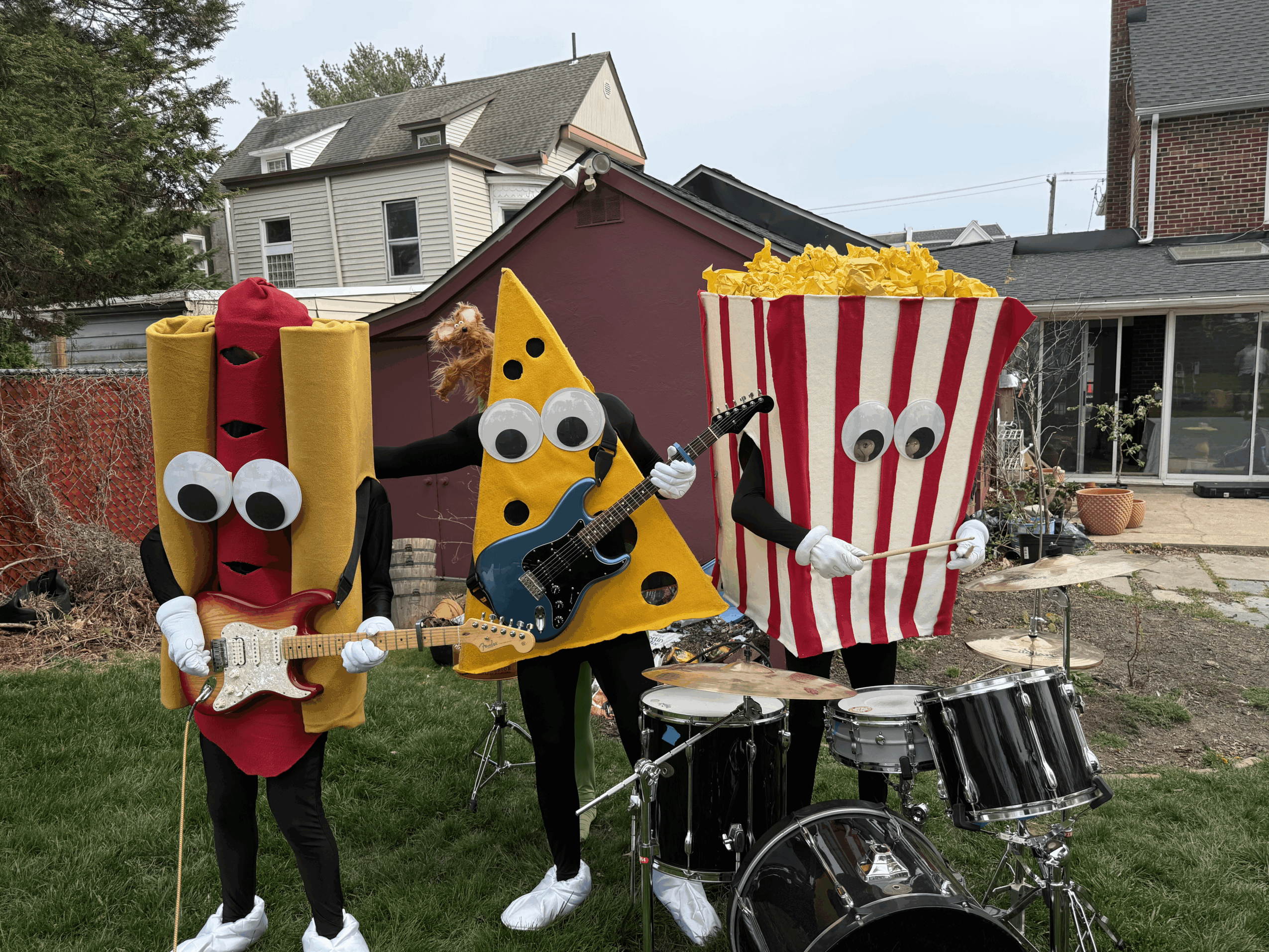 Three people in costumes resembling food items: hot dog, slice of pizza, and popcorn, playing musical instruments outdoors.