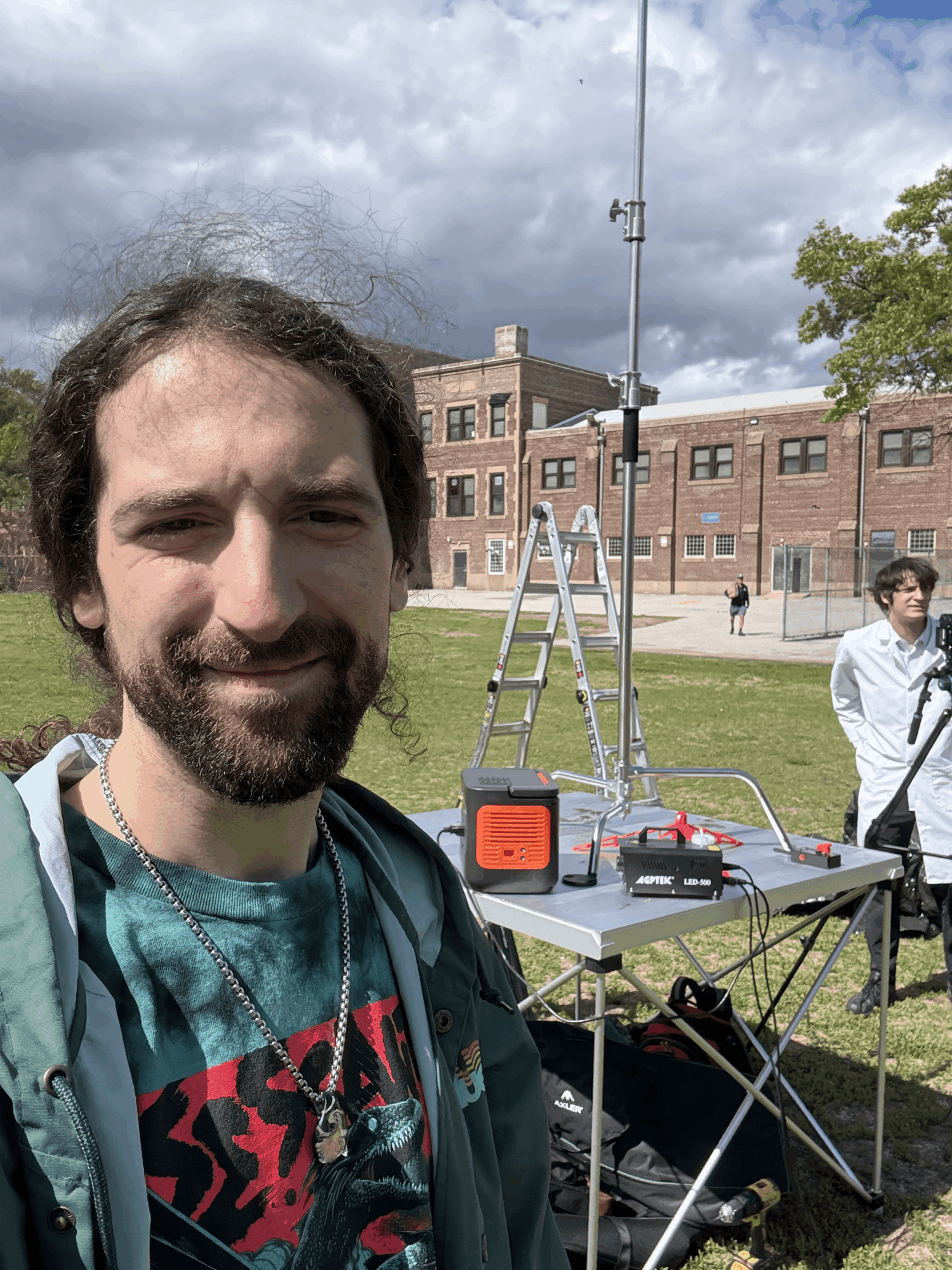 Man with beard and long hair smiling outdoors, with a table and equipment behind him, in front of a brick building.