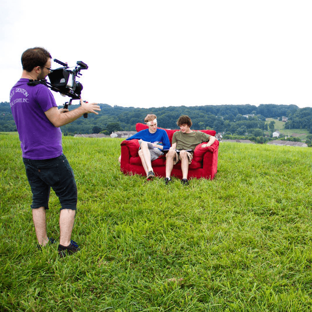 Two boys sit on a red couch in a grassy field, being filmed by a person with a camera, with a landscape background.
