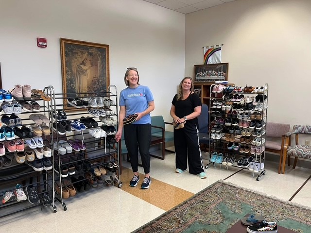 Two women stand in a room with shoe racks, smiling, with a painting on the wall and a rug on the floor.