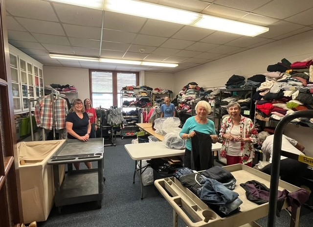 People sorting clothes in a room with shelves of folded garments and tables, window in the background.