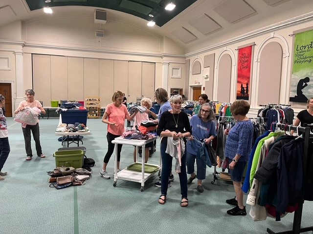 People browsing clothing racks and tables in a well-lit indoor store or market space with white walls and decorative arches.