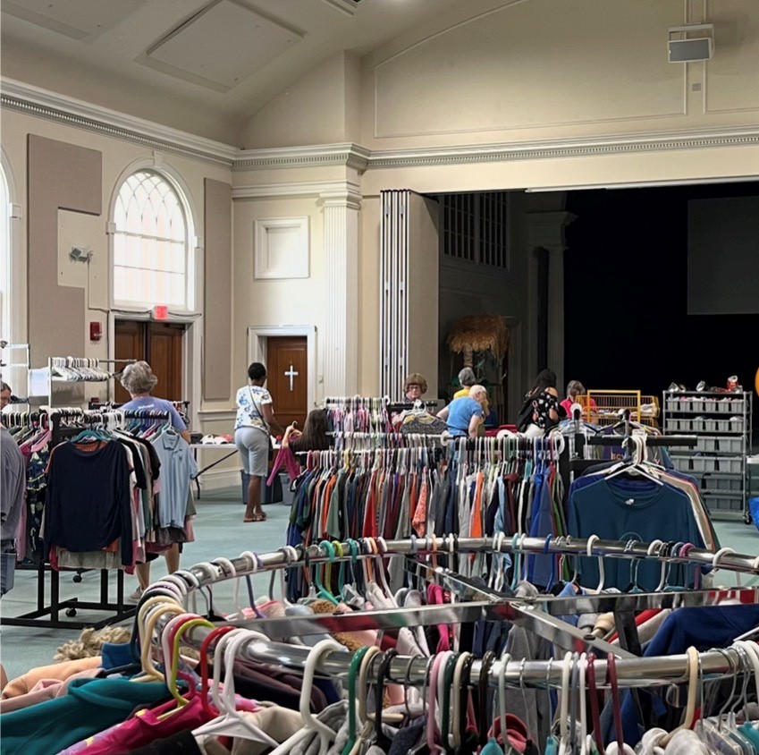 People browse clothing racks inside a spacious room with high ceilings and large windows.