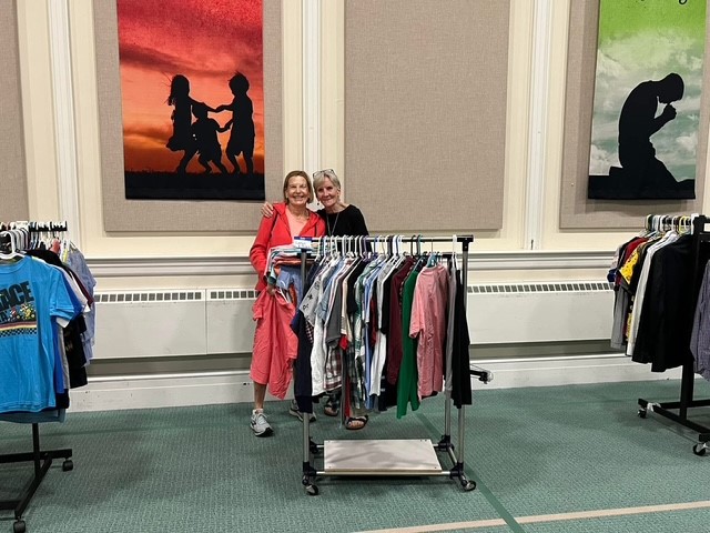 Two women stand behind a clothing rack in a store, smiling, with framed silhouette artwork on the wall behind them.