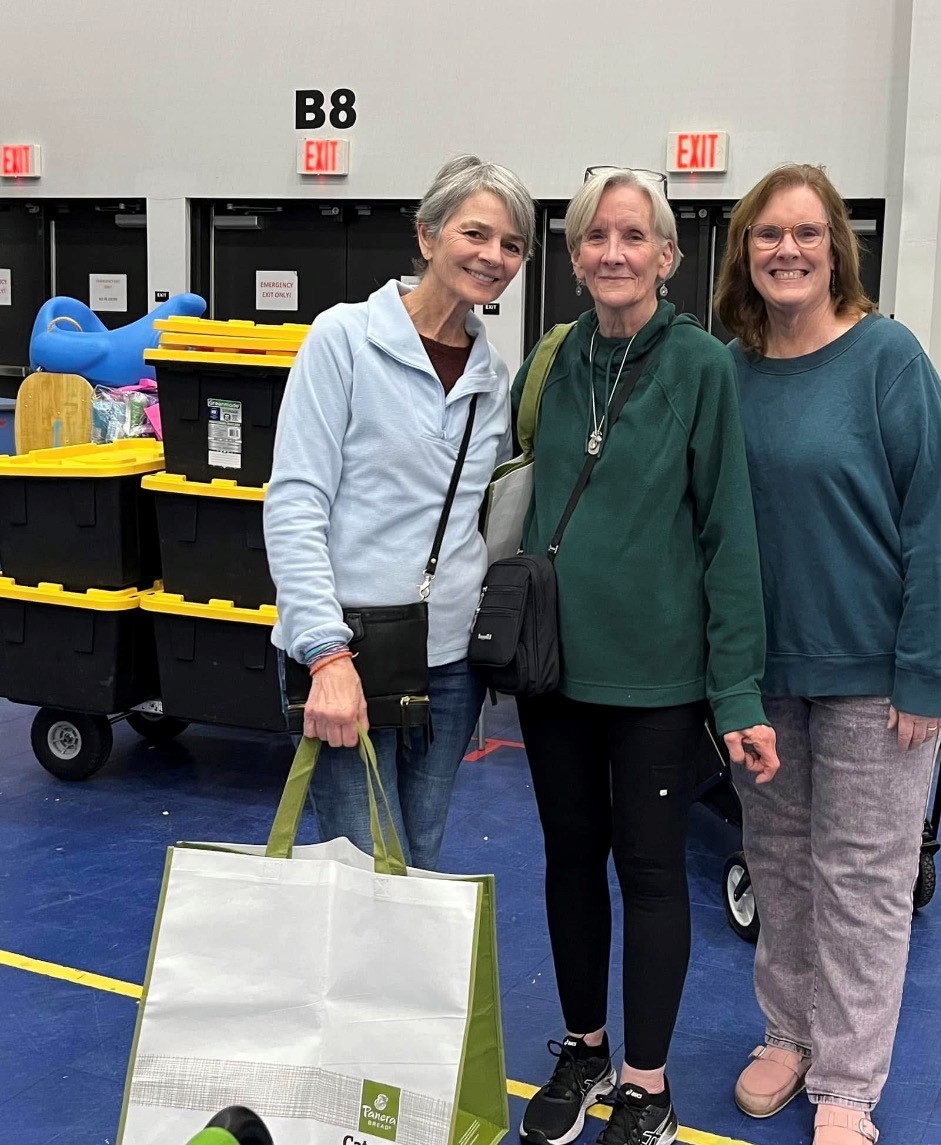 Three women standing together indoors, smiling, with storage bins and exit signs in the background.