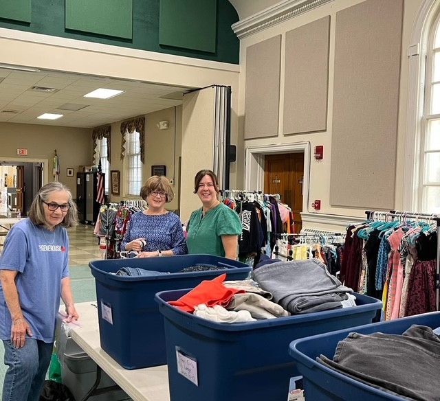 Three women standing behind large blue bins filled with clothes in a room with racks of clothing and high ceilings.