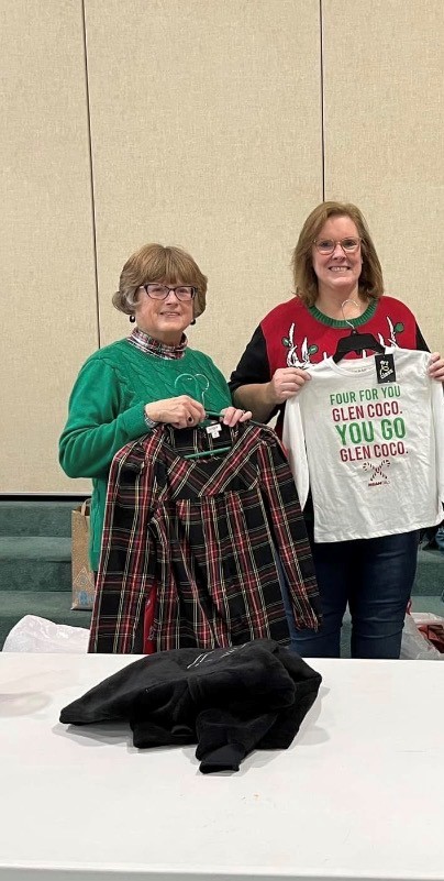 Two women standing indoors, one holding a plaid garment and a bag, with a table in front. Both are smiling.
