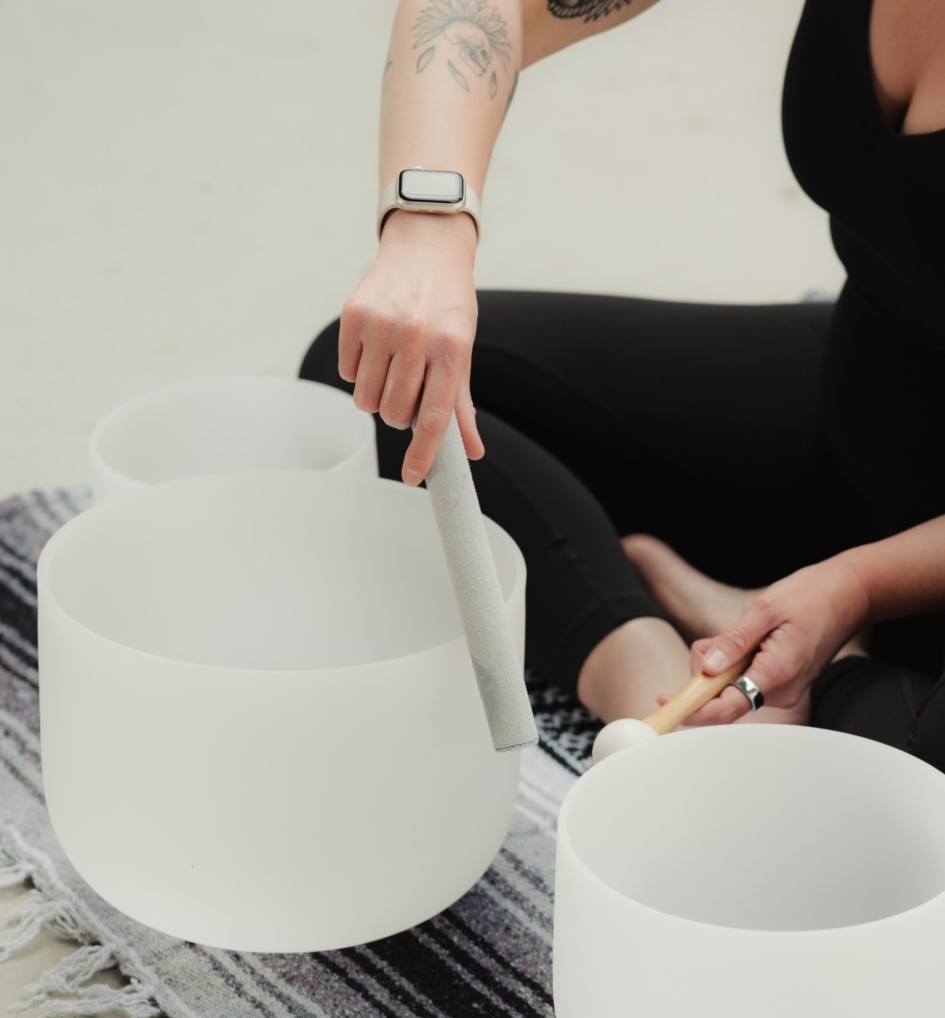 Person playing crystal singing bowls, wearing black clothing, with tattoos and a smartwatch, sitting on a striped rug.