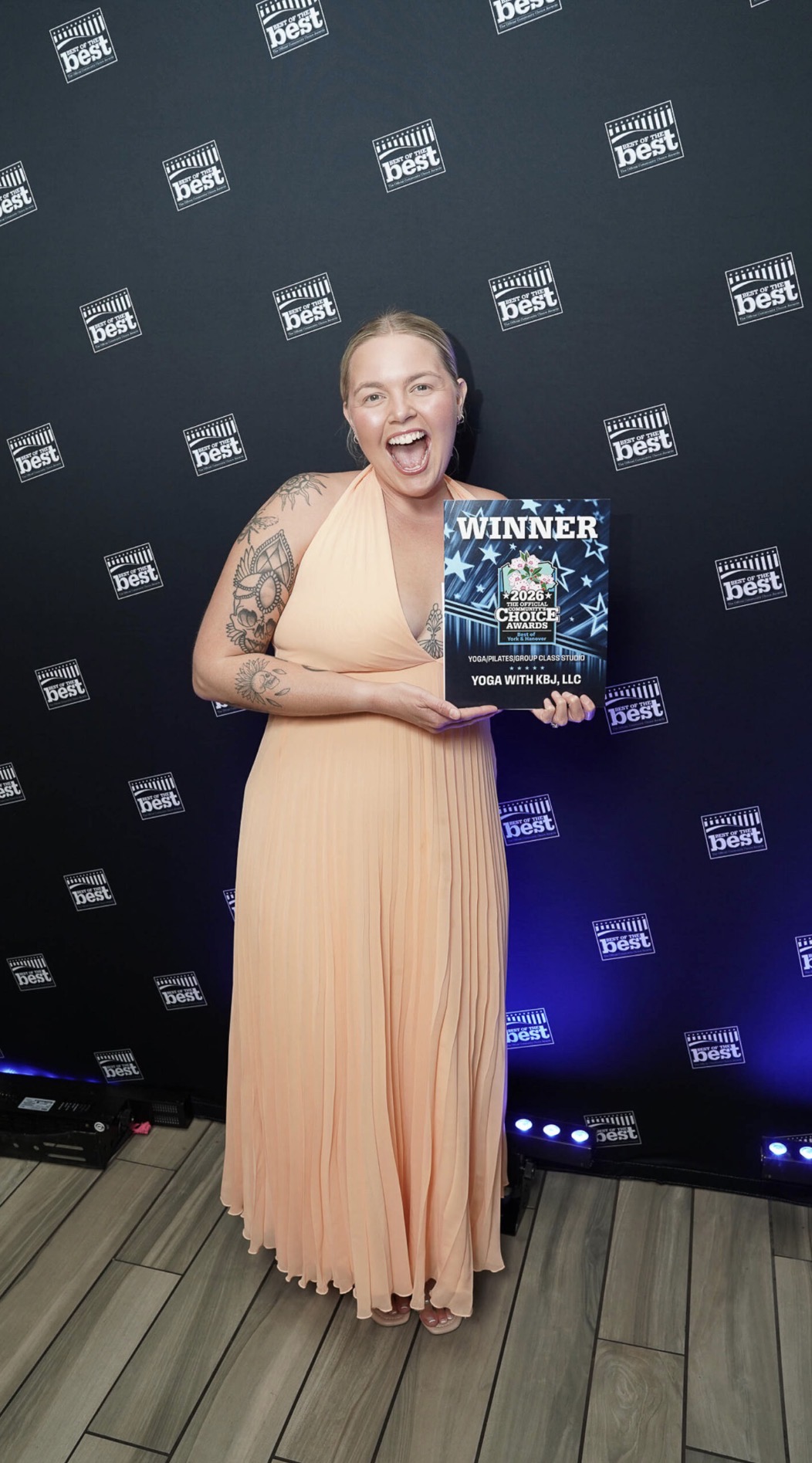 Woman in peach dress holding a winner certificate, smiling, standing in front of a black backdrop with logos.