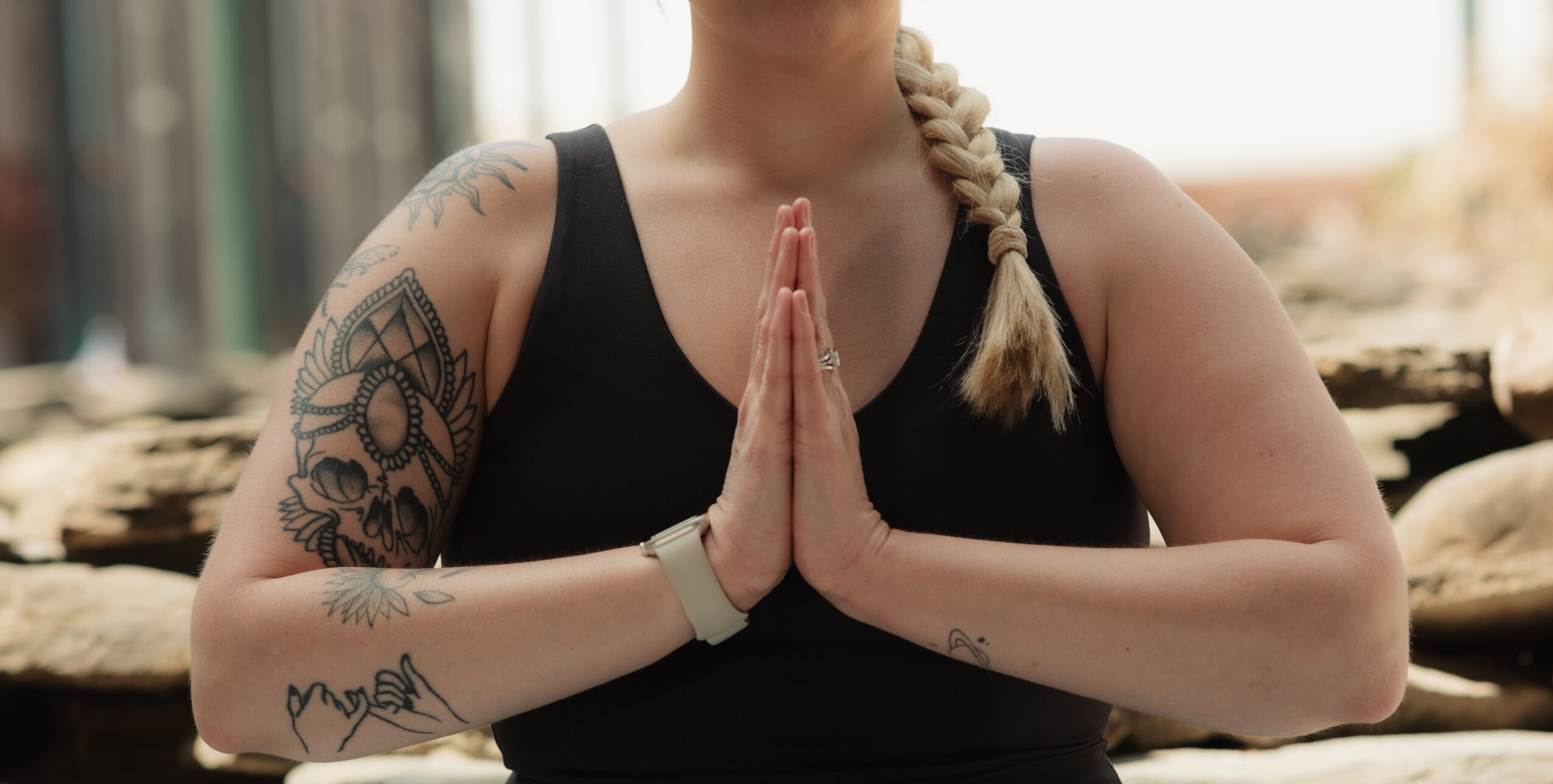 Person with tattoos on arms, hands pressed together in prayer pose, wearing a black tank top, outdoors.