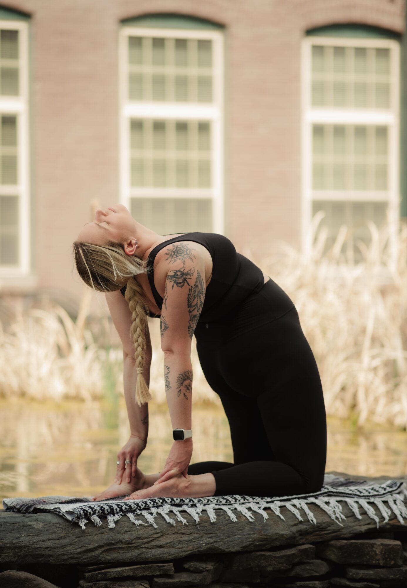 Woman with tattoos in black dress kneeling on a patterned blanket outdoors, with a building and tall grass in background.
