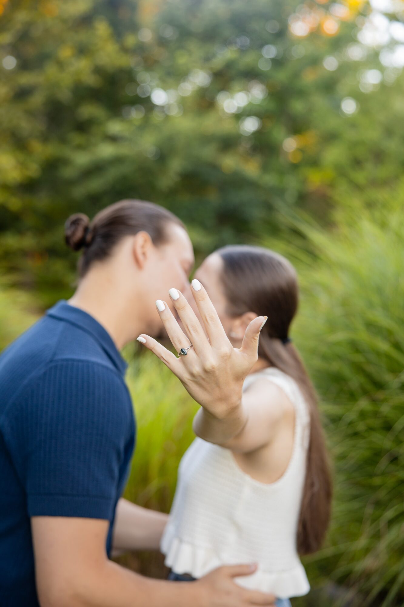 Two women face each other outdoors, one showing her hand with a ring, surrounded by greenery.