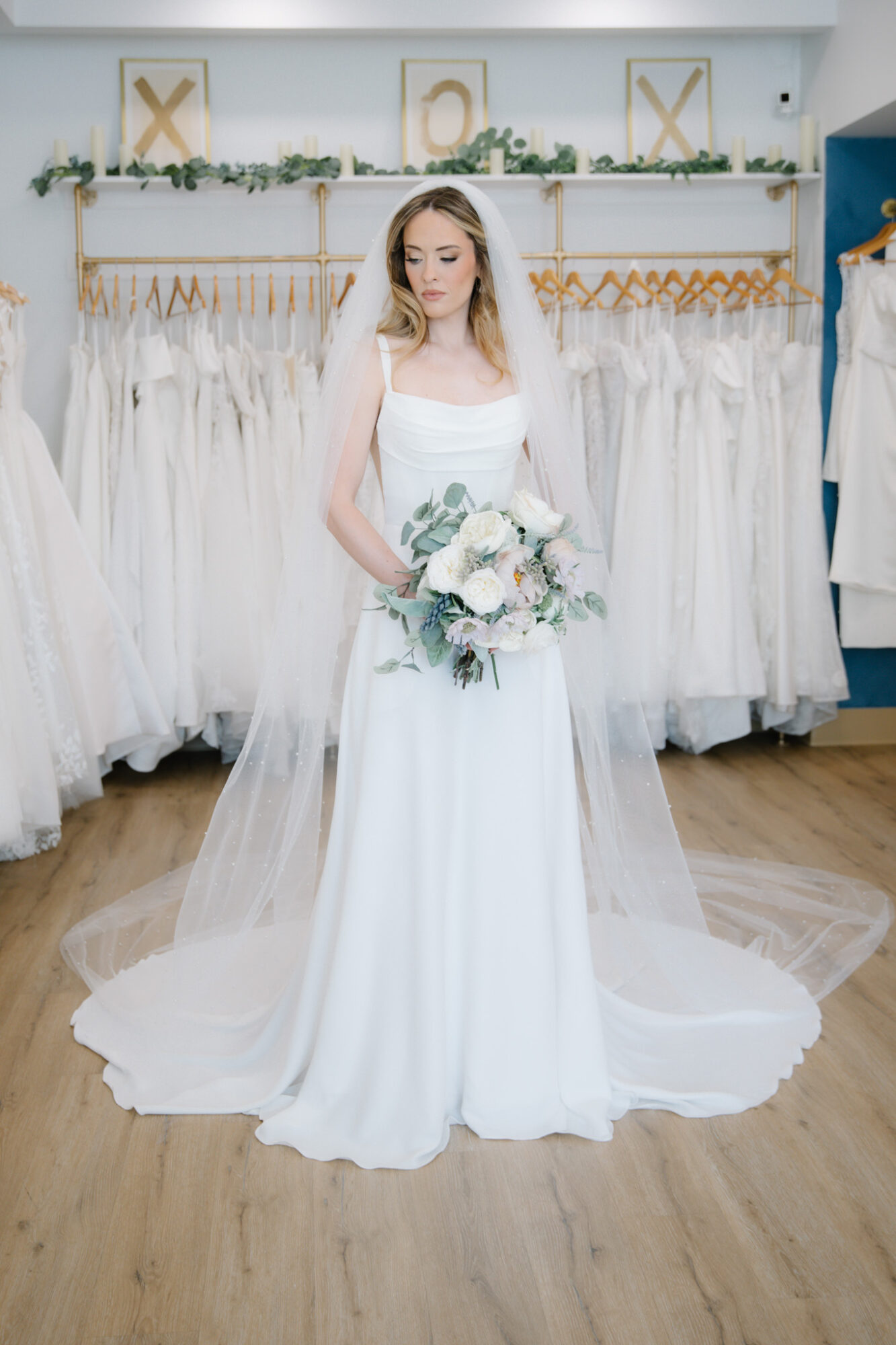 Bride in a white wedding dress holding a bouquet, standing in a bridal shop with dresses on racks behind her.