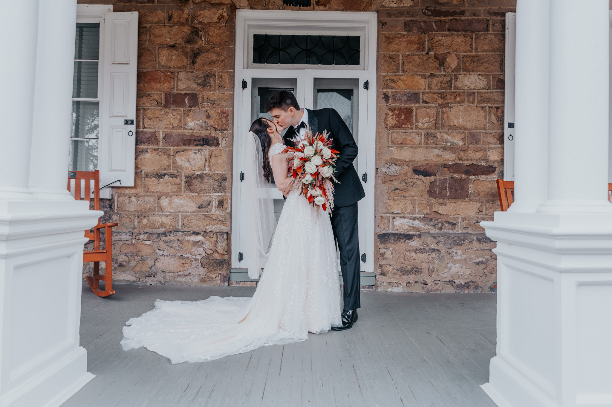 Bride and groom kissing in front of a brick building, bride holding a bouquet, wedding dress flowing behind, columns on sides.
