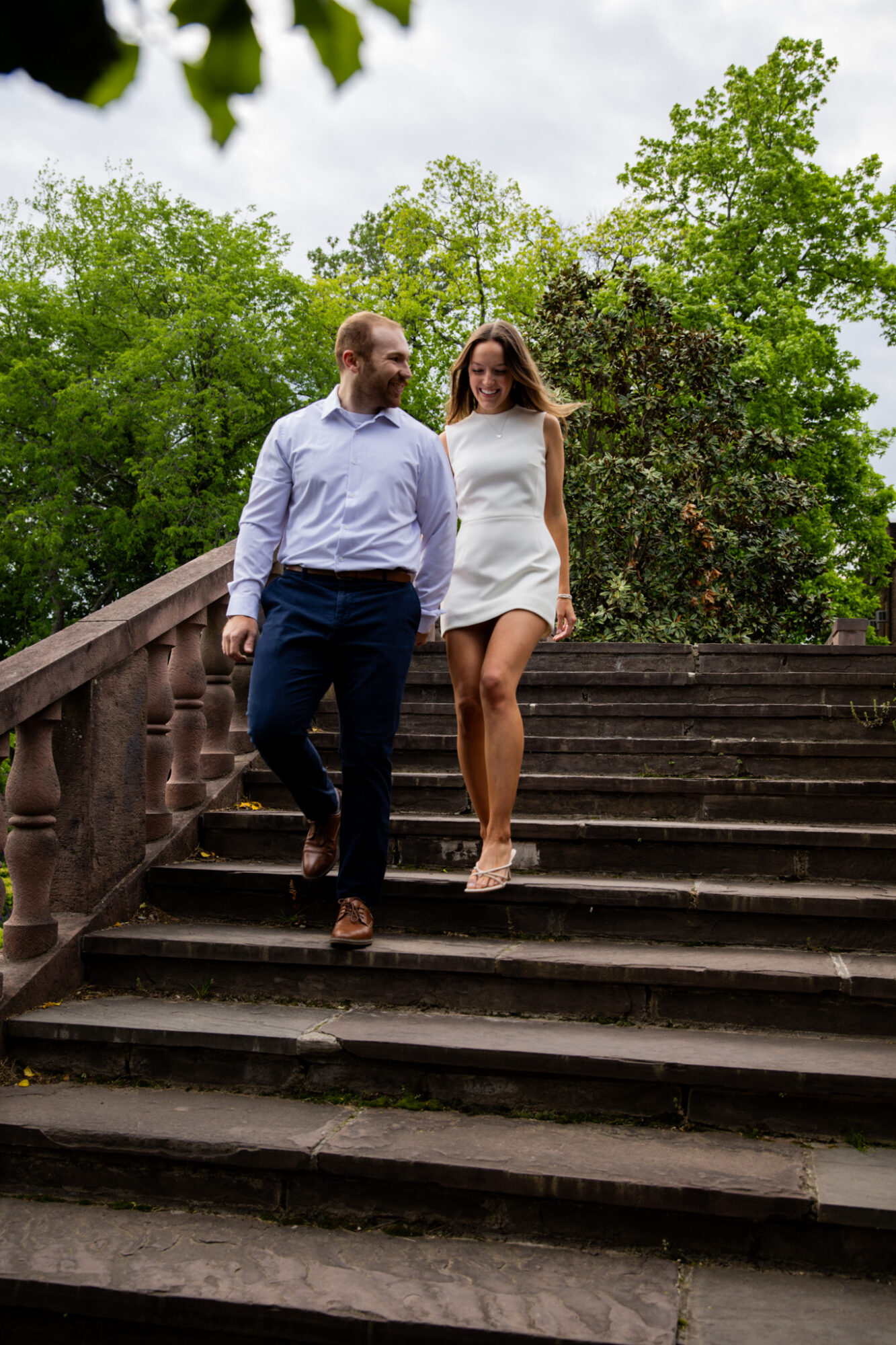 A man and woman walk down outdoor stairs surrounded by green trees, holding hands and smiling.