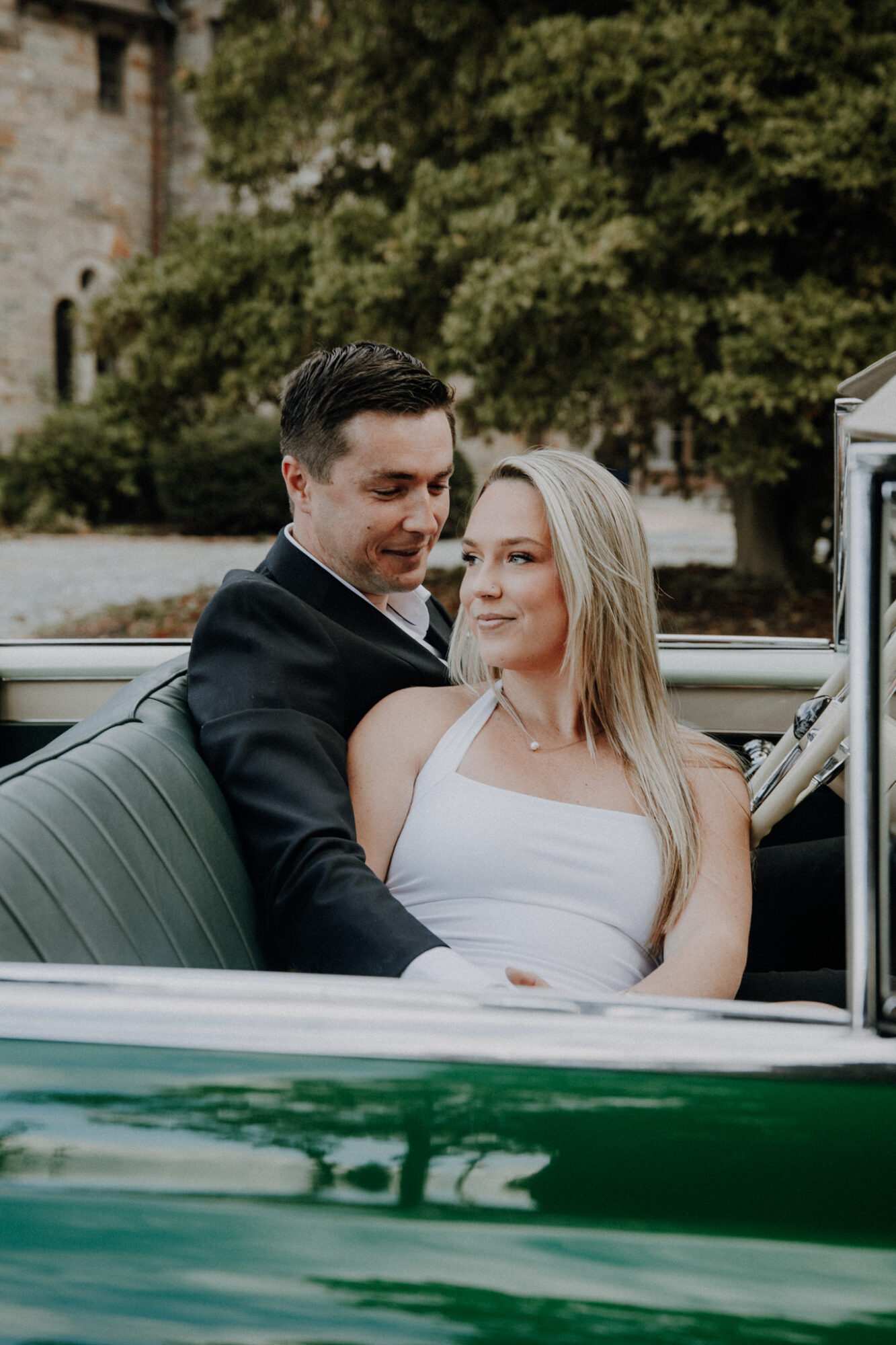 A man and woman sit in a vintage green car, smiling and looking at each other, outdoors with trees and a building in background.