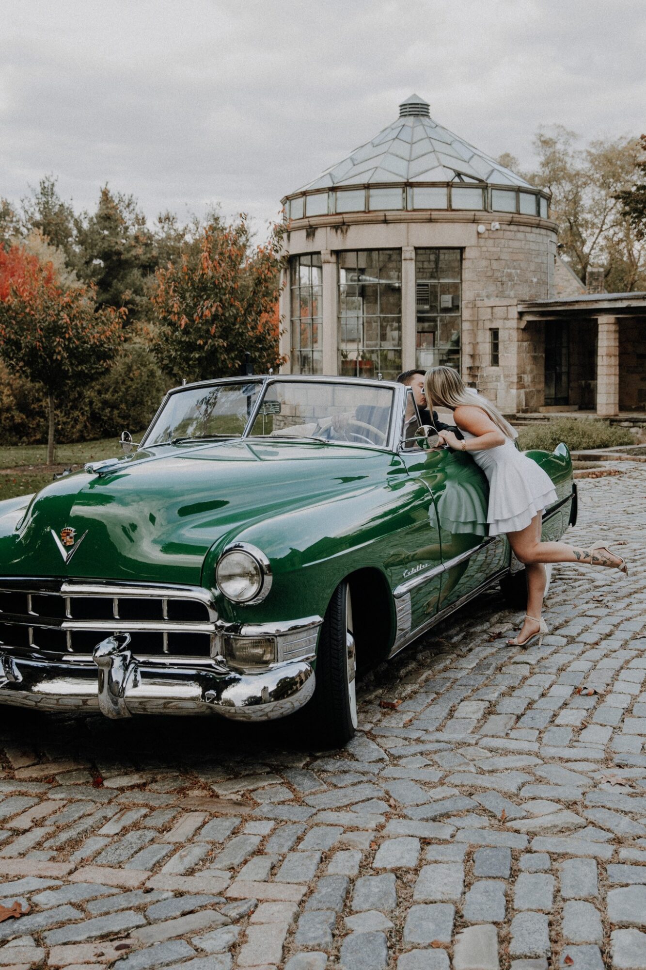 Person leaning on a vintage green car parked on cobblestone, with a round stone building and trees in background.