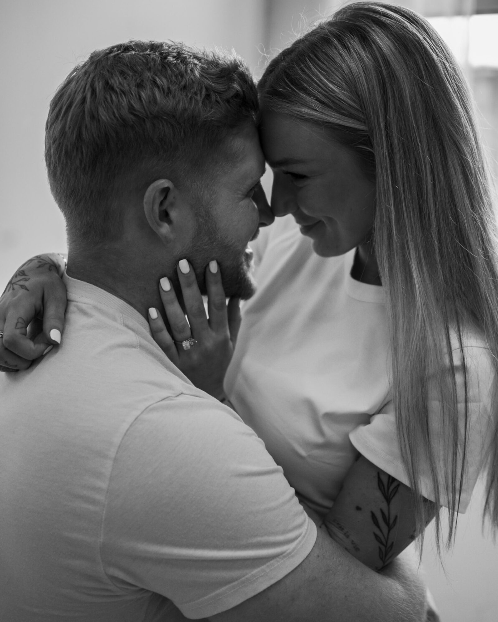 A couple faces close, touching foreheads, smiling, with one person's hand on the other's shoulder, in a cozy setting.