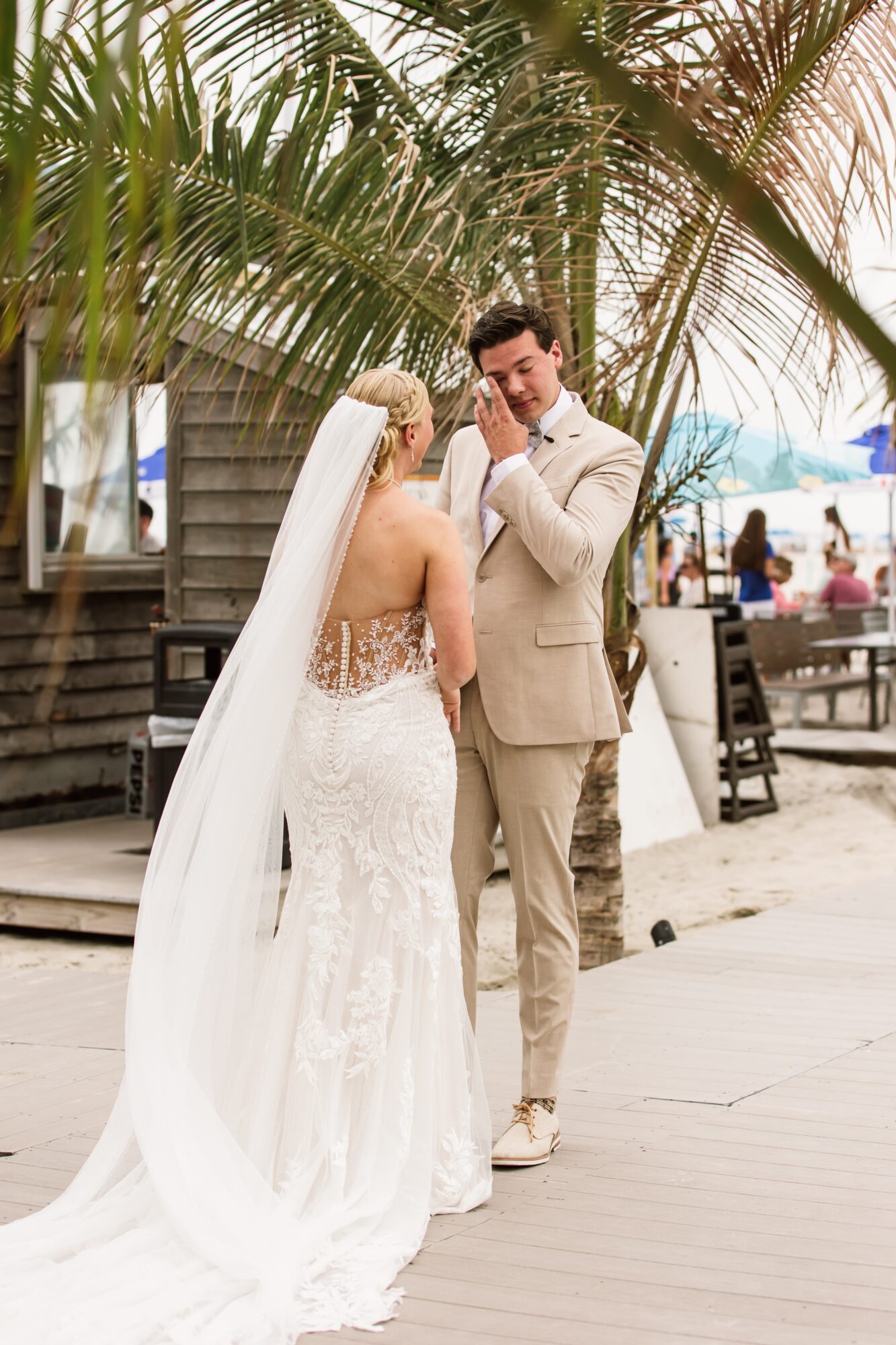 Bride and groom stand close together outdoors, surrounded by palm trees and wooden benches, with people in the background.