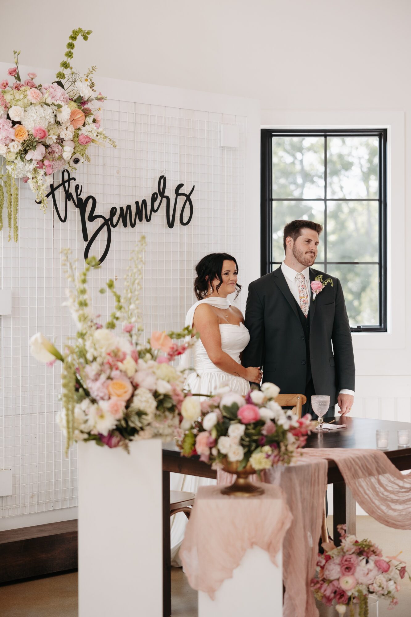Bride and groom standing indoors near a window, with floral arrangements and a sign reading 'The Benners'.