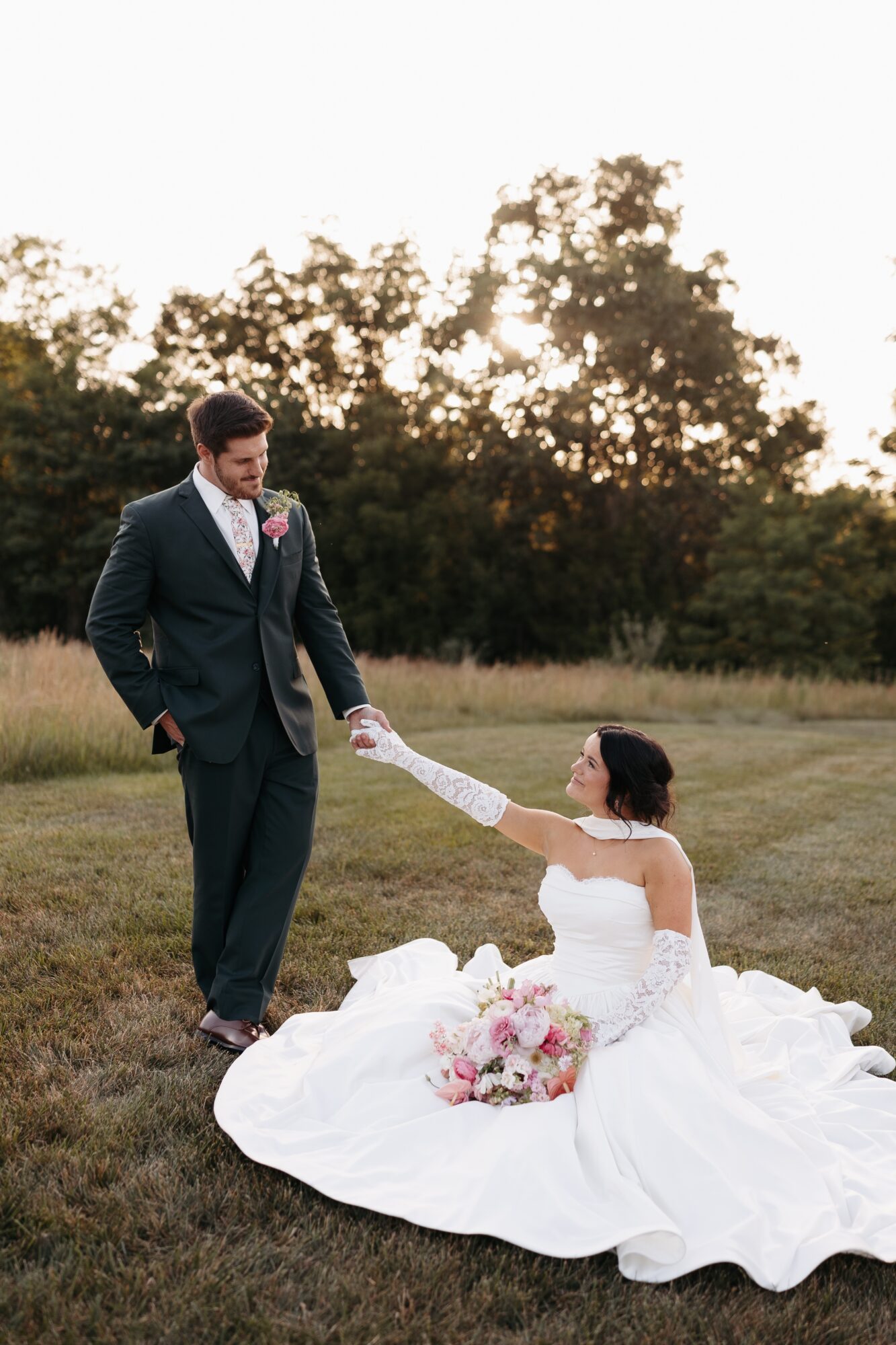 Bride sitting on grass in wedding dress, reaching out to groom standing nearby, with trees and sky in background.