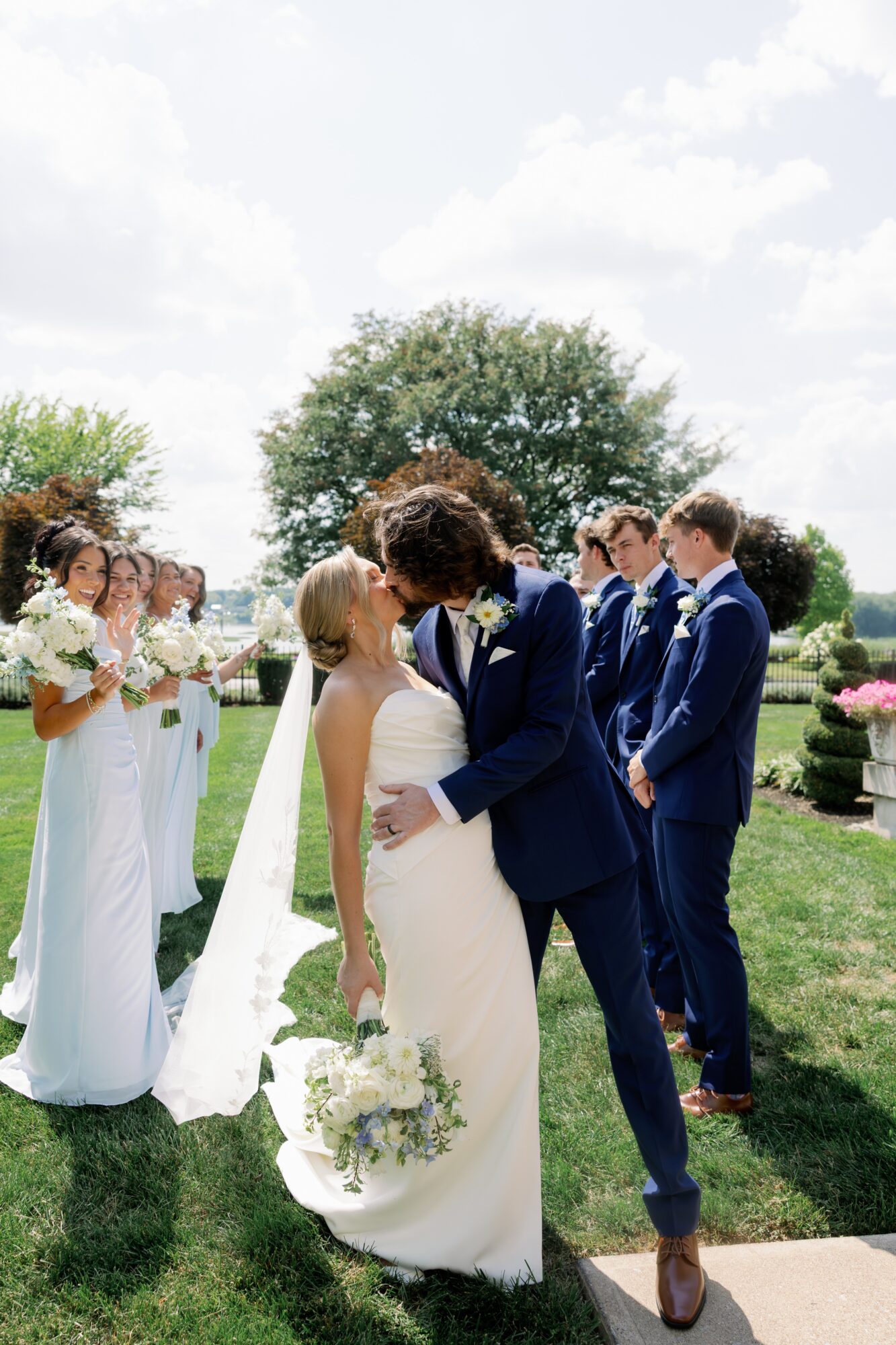 Couple kissing at wedding ceremony outdoors, surrounded by bridesmaids and groomsmen, with trees and sky in background.