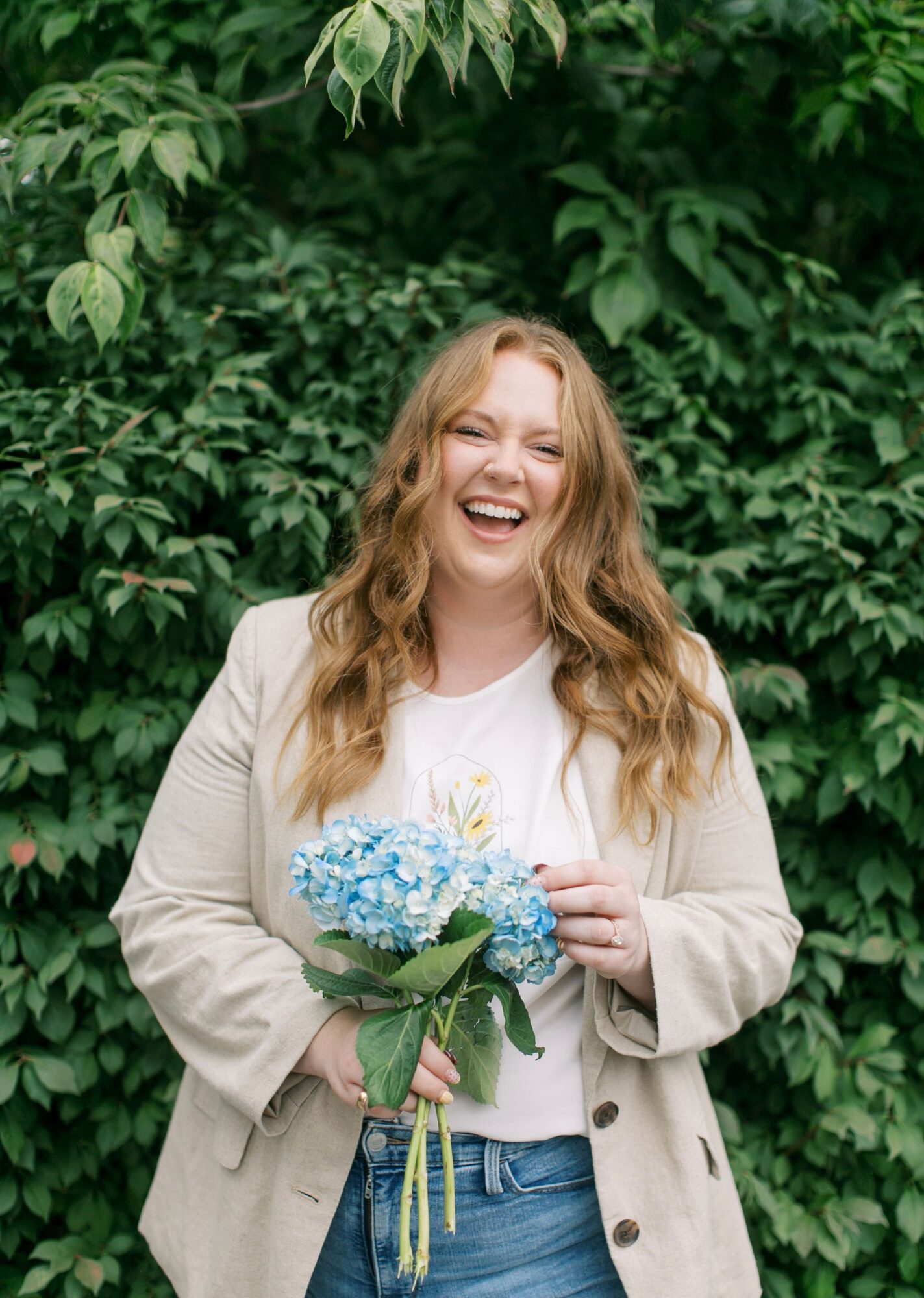Woman with long wavy hair smiling, holding a bouquet of blue flowers, standing in front of green leafy background.