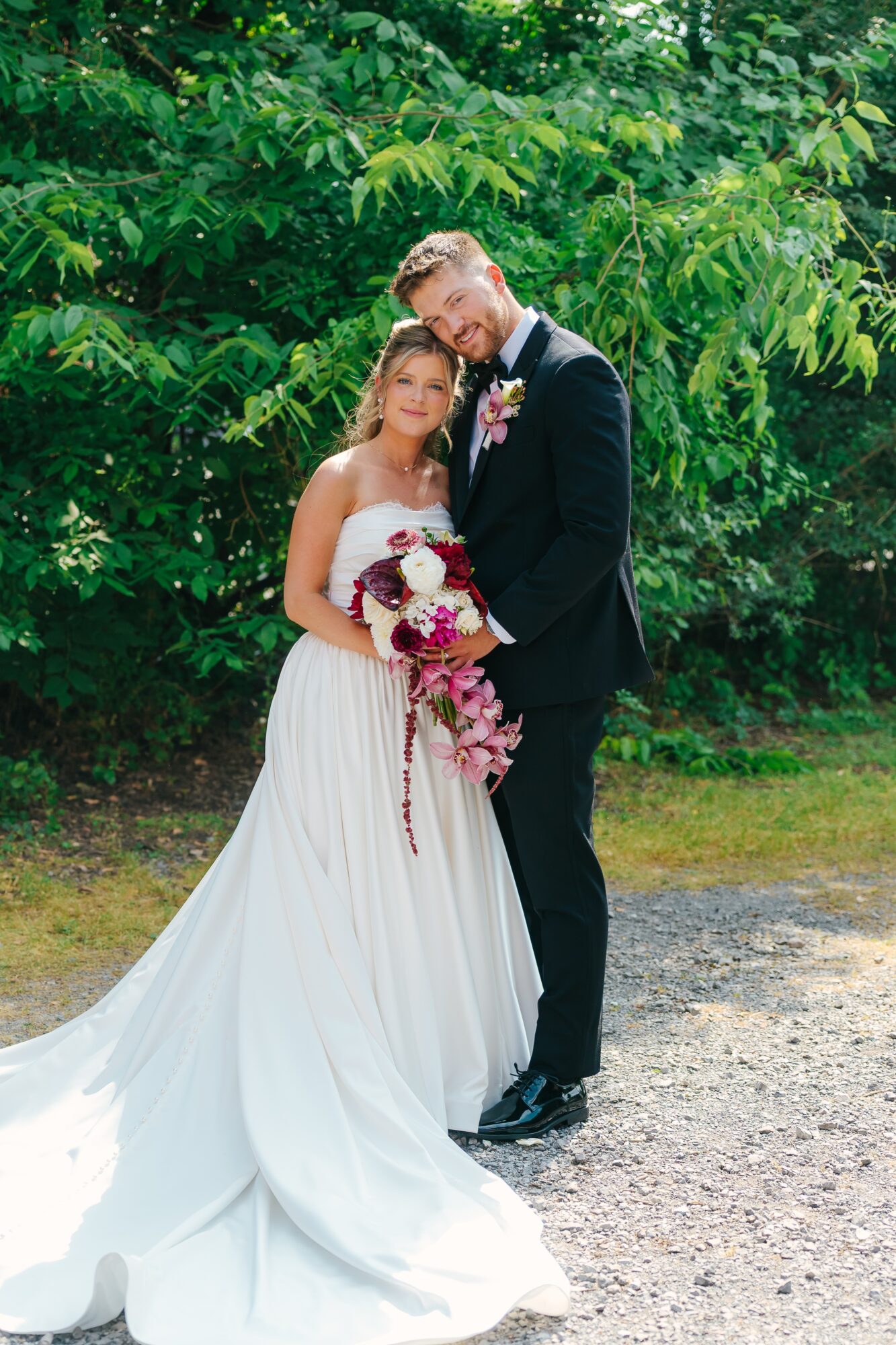 Bride and groom standing outdoors, bride holding a bouquet, surrounded by green trees, both smiling.
