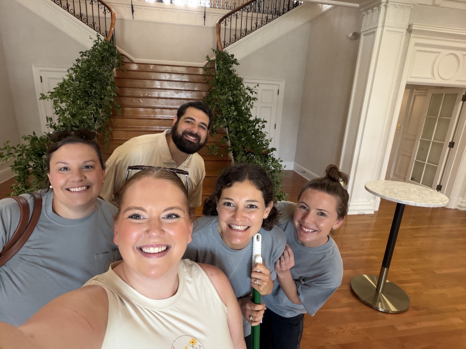 Group of five smiling people taking a selfie indoors near a staircase with greenery, wooden floors, and a round table.