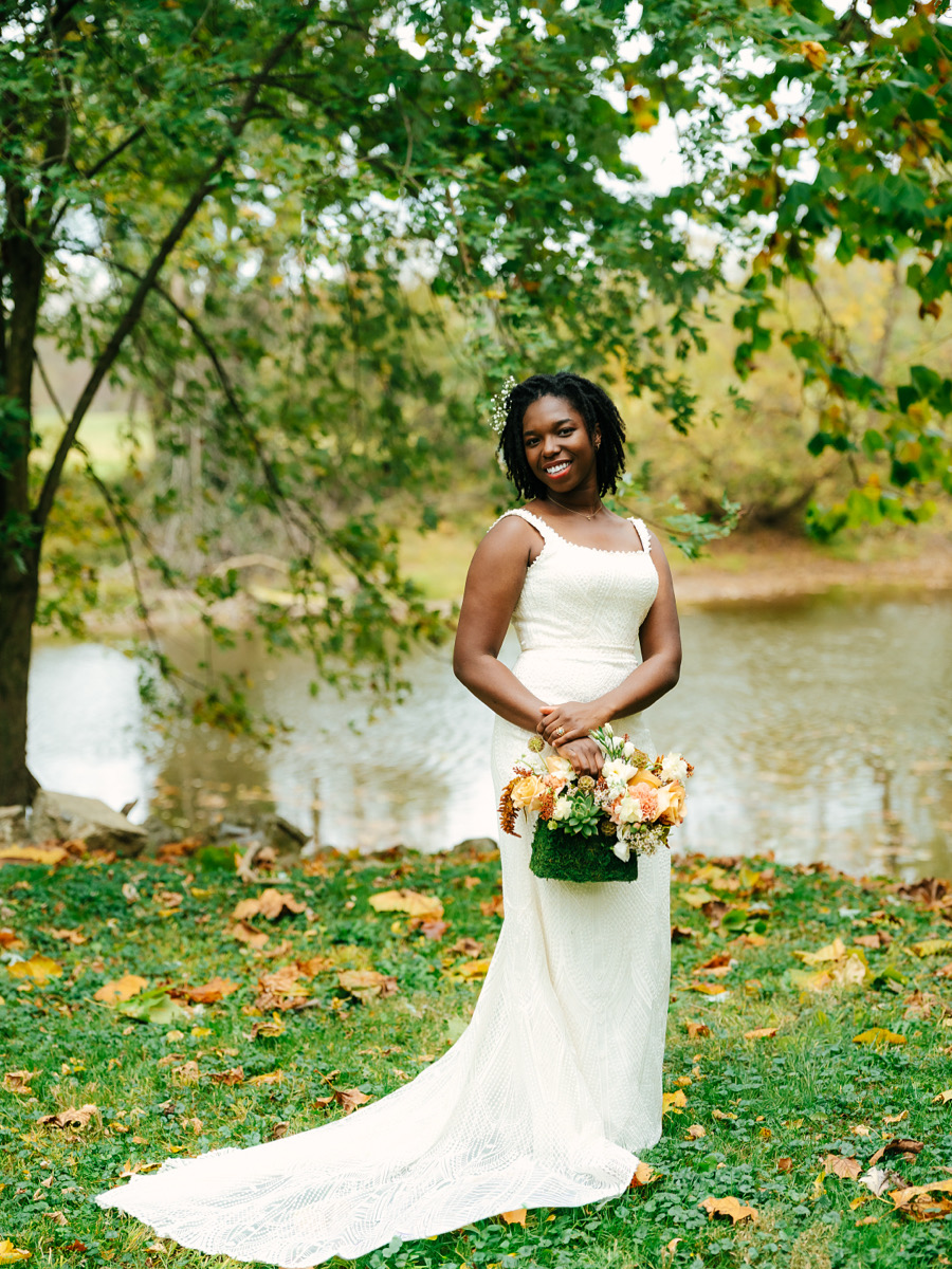 A woman in a white dress holding a bouquet stands outdoors near a river, surrounded by trees with green leaves.