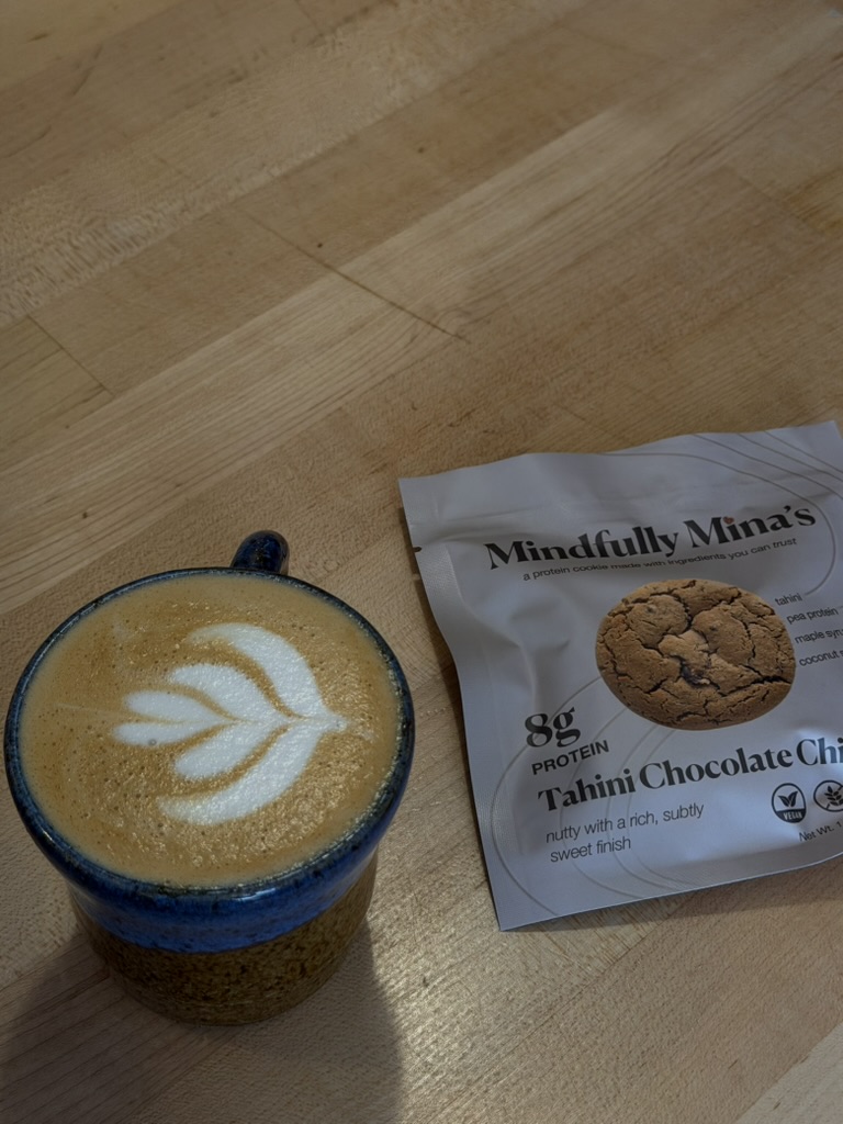 A glass of coffee with latte art next to a packaged cookie on a wooden surface.