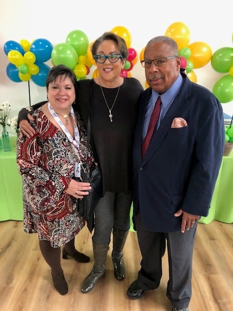 Three people standing together in front of colorful balloons, smiling, indoors with a green tablecloth, wooden floor.