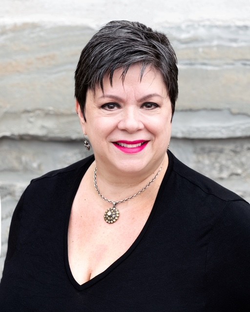 Woman with short dark hair smiling, wearing a black top and a necklace, standing outdoors with a stone background.