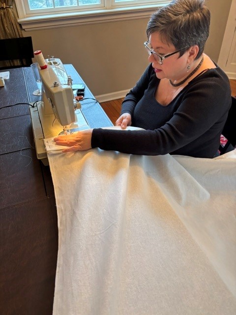 Woman sewing fabric on a sewing machine at a table, with a window in the background.