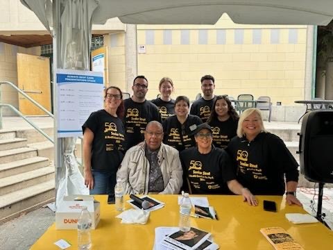 Group of nine people, some wearing black shirts, gathered around a yellow table outdoors, with stairs and a building in the background.
