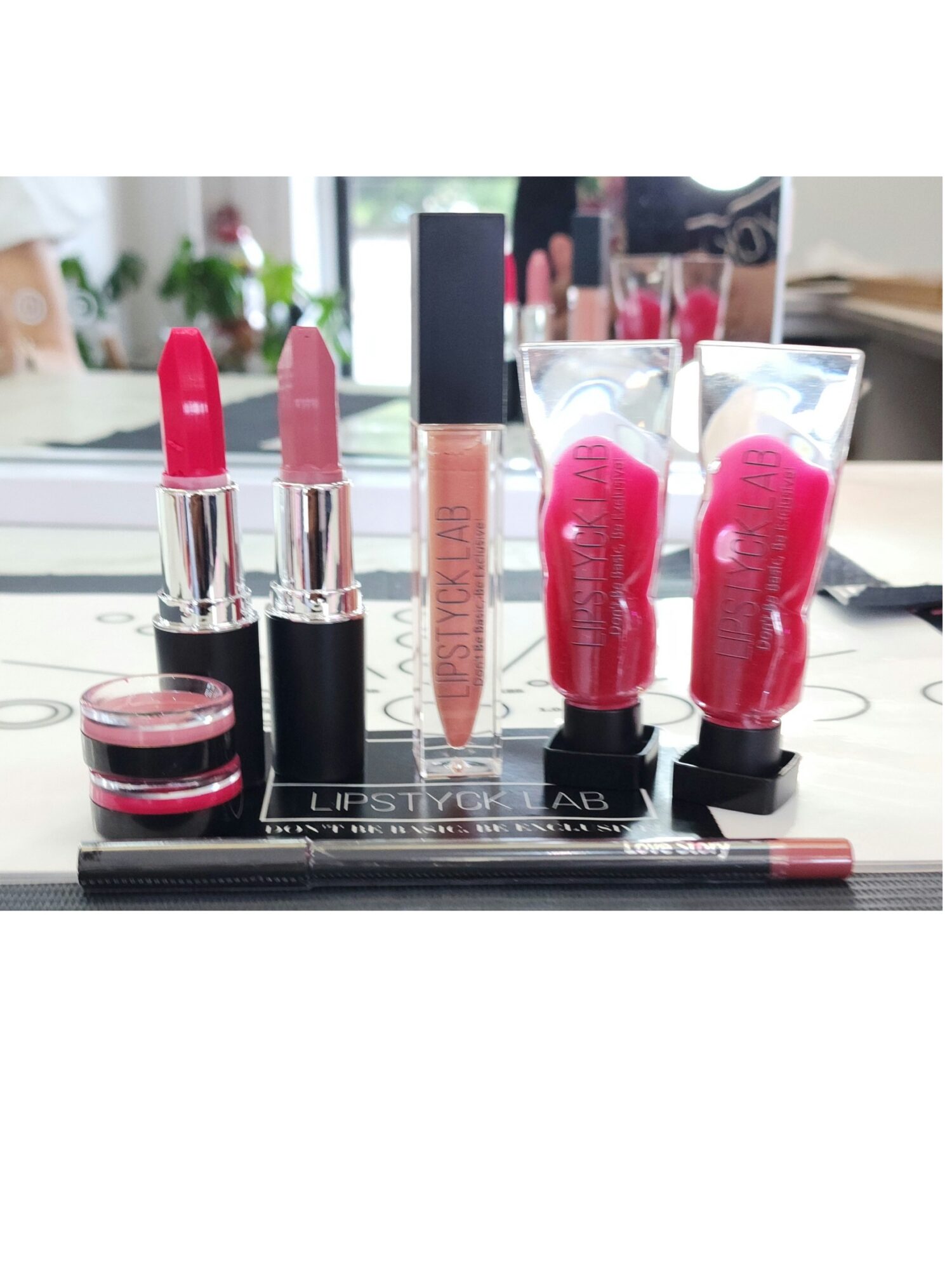 Various lipsticks, lip gloss, and lip balms arranged on a black tray with a blurred background of a window and plants.